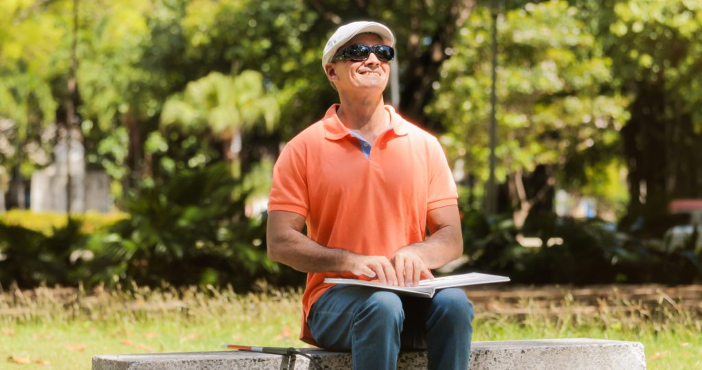 Handicapped People With Disability Blind Man Reading Braille Book Hispanic blind man, latino people with disability, handicapped person and everyday life. Visually impaired man reading book with hands, touching page written in Braille language with fingers.