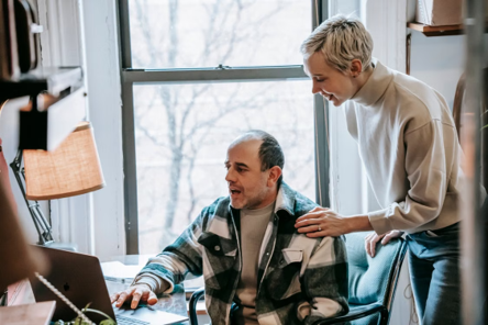 A man in a plad shirt sits a computer while a woman with short hair looks on from behind him. Stock photo by Gary Barnes via Pexels. 