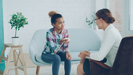 A young African-American woman, sitting on a sofa talks to a white woman seated in a chair across from her. Stock photo by Vitaly Gariev via Pexels