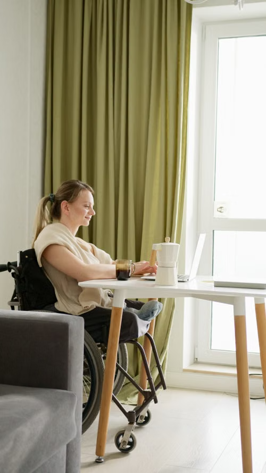 A woman in a wheelchair working on a laptop in a bright indoor space with green curtains.