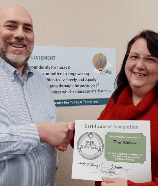 A bald white hispanic male witj a beard wearing a blue dress shirt shakes hands with a white woman with black hair wearing glasses and a red sweater, the woman is displaying a certificate