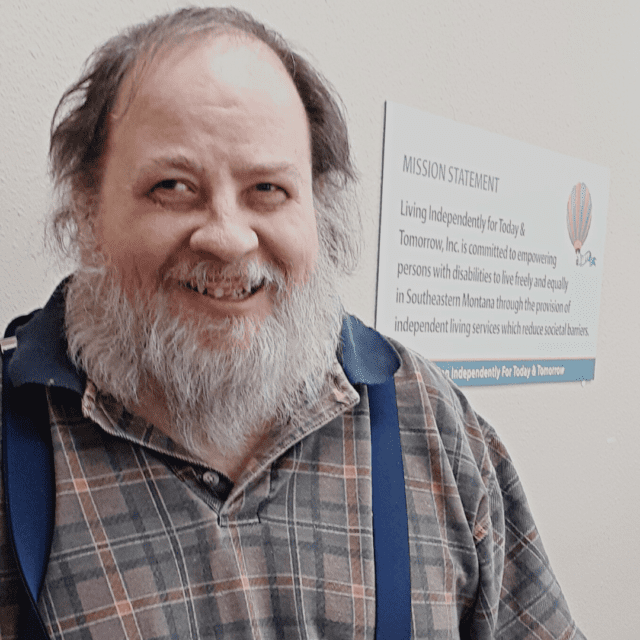 white male with reccding black hair and a grey beard wearing a brown & blue plaid shirt with suspenders